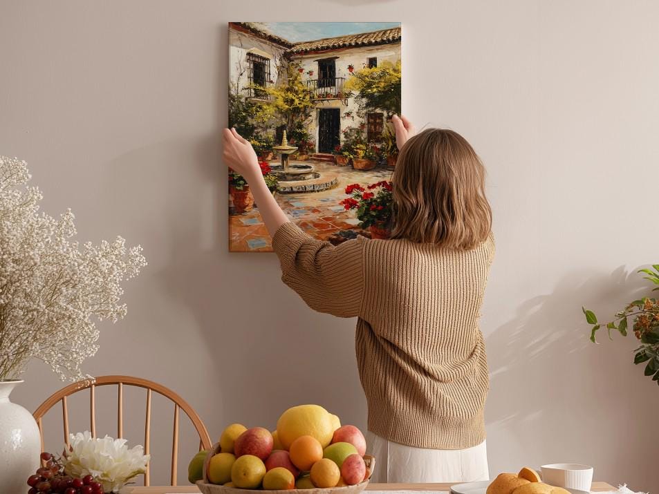 A woman hangs a framed picture of a Spanish-style house on a wall in a cozy, homely setting.