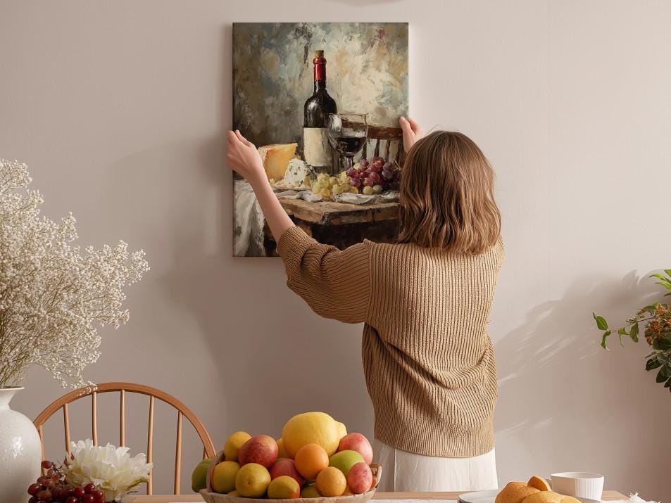 A woman hangs a painting of a table with wine, cheese, and fruit on a wall in a cozy, homely setting.