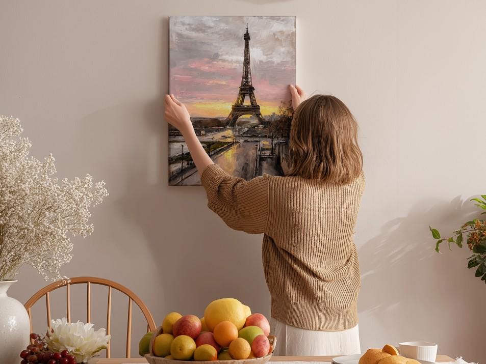 A woman hangs a framed picture of the Eiffel Tower on a wall in a room with a dining table, chairs, and various fruits.