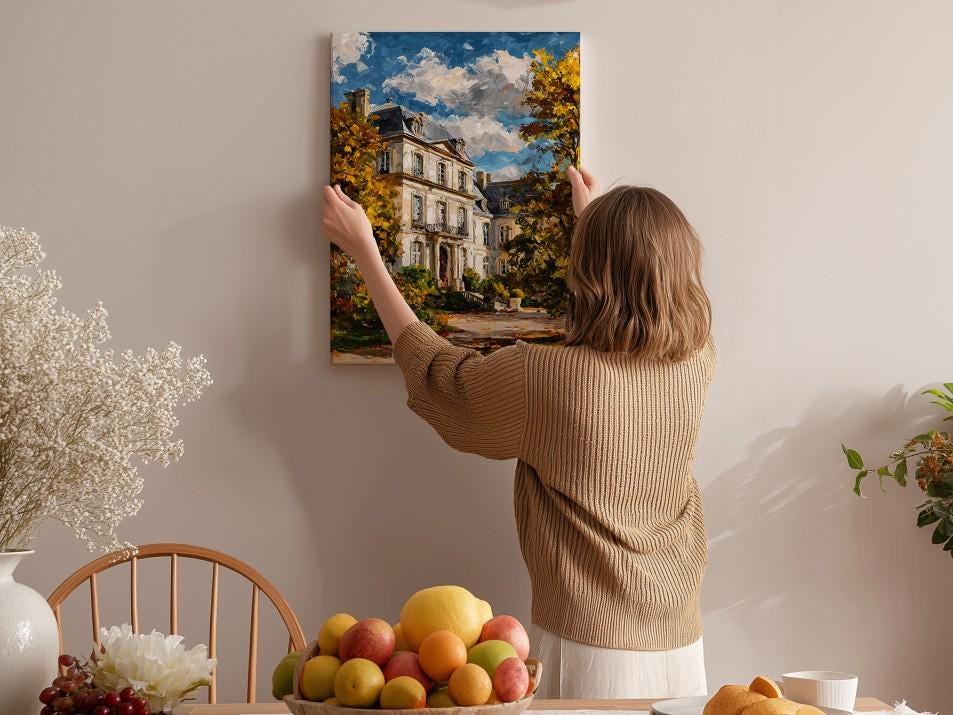 A woman hangs a painting of a house on a wall in a room with a dining table, chairs, and various fruits and flowers.