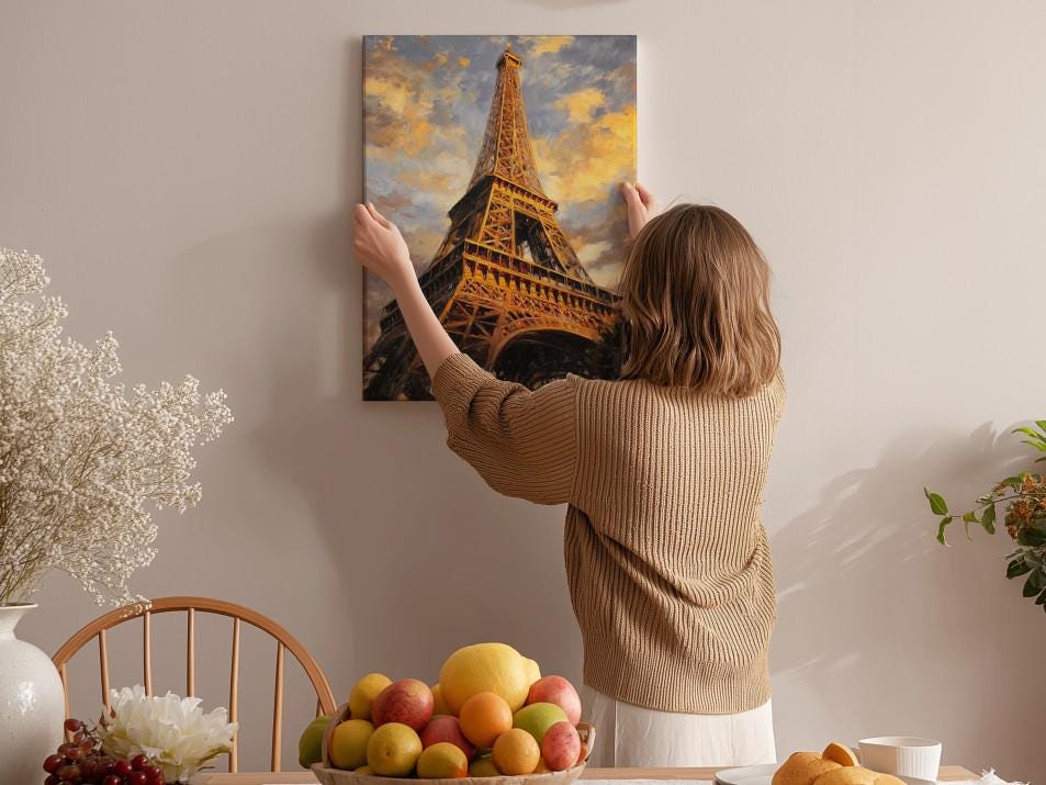 A woman hangs a framed picture of the Eiffel Tower on a wall in a room with a dining table, chairs, and various decorative items.