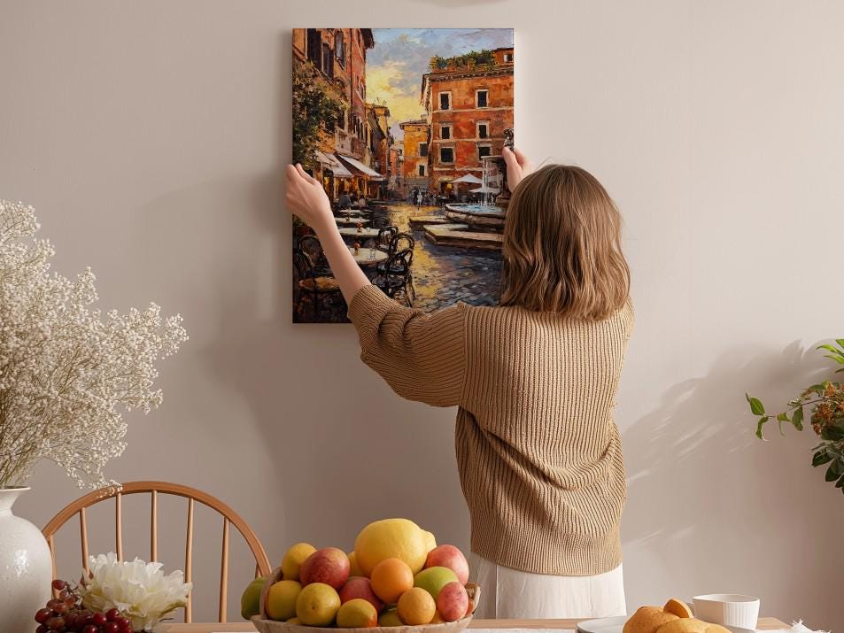 A woman hangs a painting of a cityscape on a wall in a cozy room with a dining table, chairs, and various decorative items.