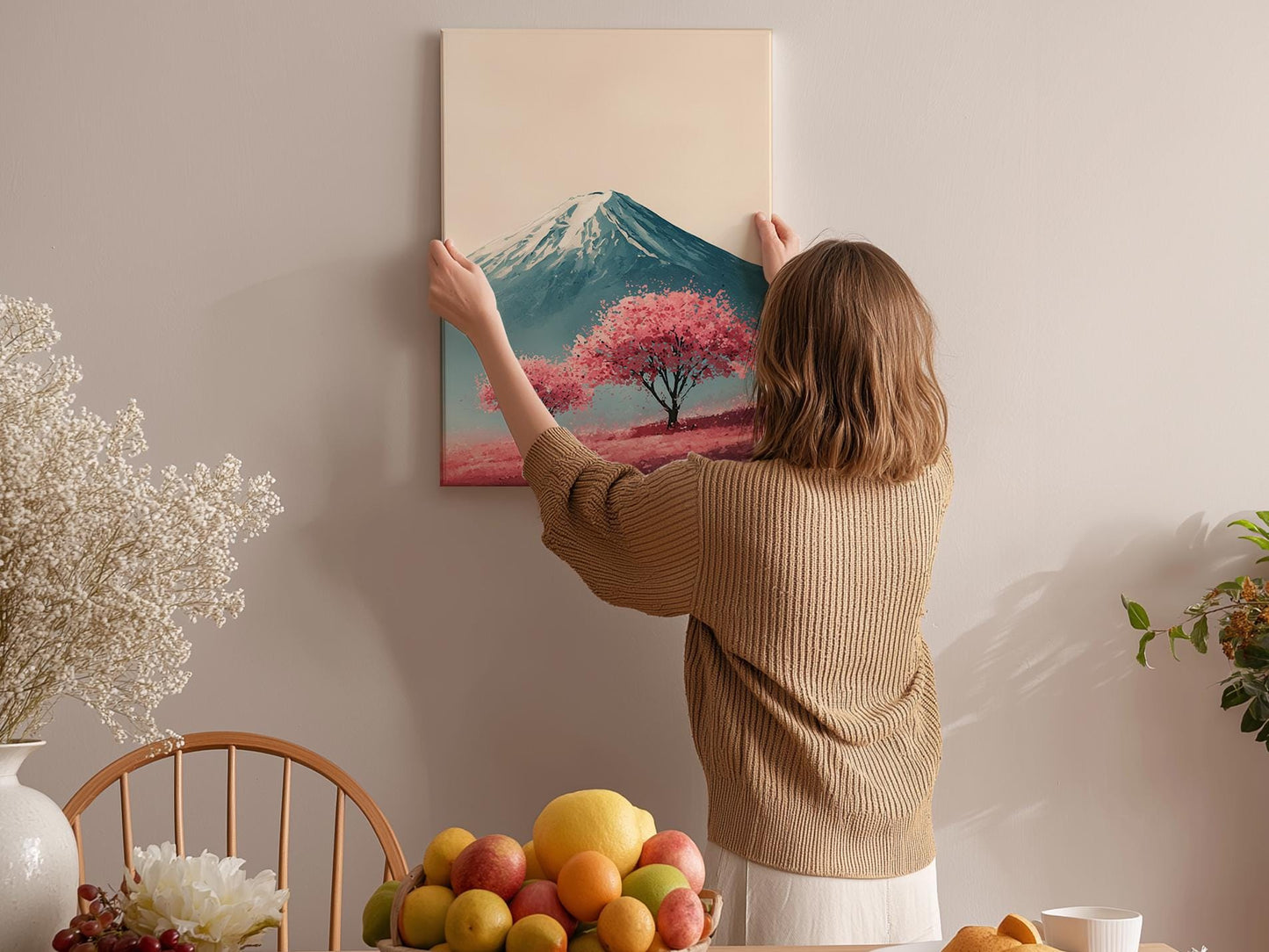 A woman hangs a framed picture of a mountain with a pink tree on a wall in a cozy room with a dining table, chairs, and various fruits.