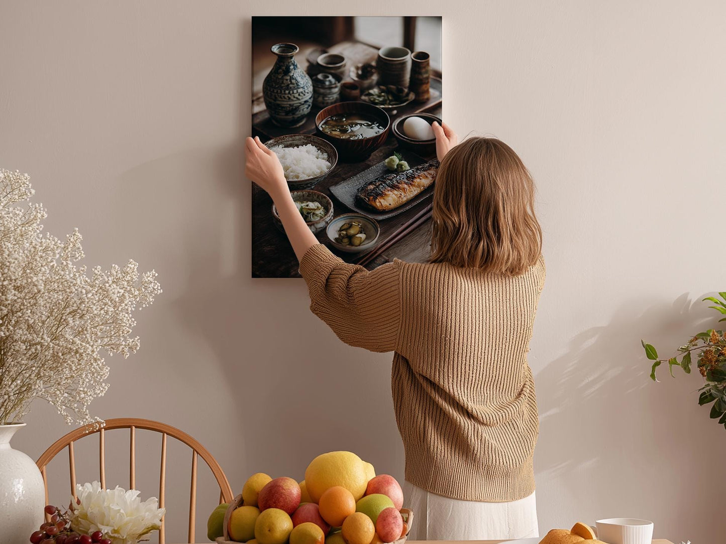 A woman hangs a framed photograph of a table with various dishes, including bowls, plates, and a vase, on a wall in a room with a dining table and chairs.