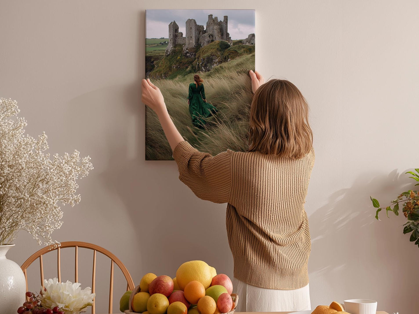 A woman hangs a framed picture of a castle on a wall in a room with a dining table, chairs, and various fruits.