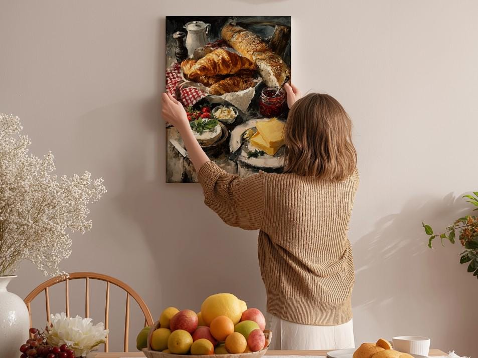 A woman hangs a framed painting of a table with various food items, including bread, fruit, and cheese, on a wall in a cozy, homely setting.