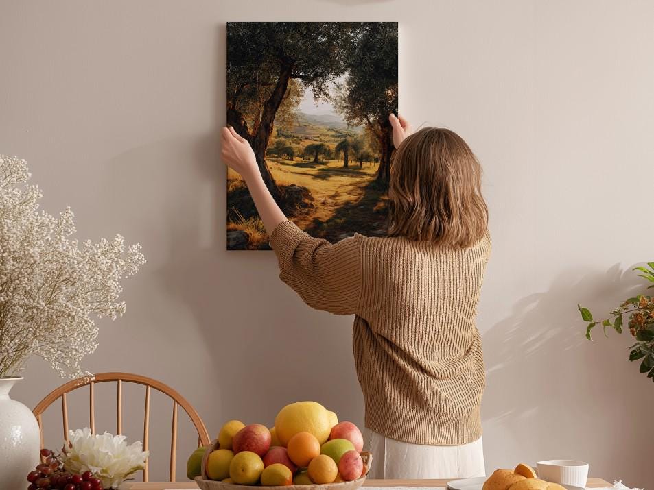 A woman hangs a framed landscape painting on a wall in a cozy room with a dining table, chairs, and various fruits.