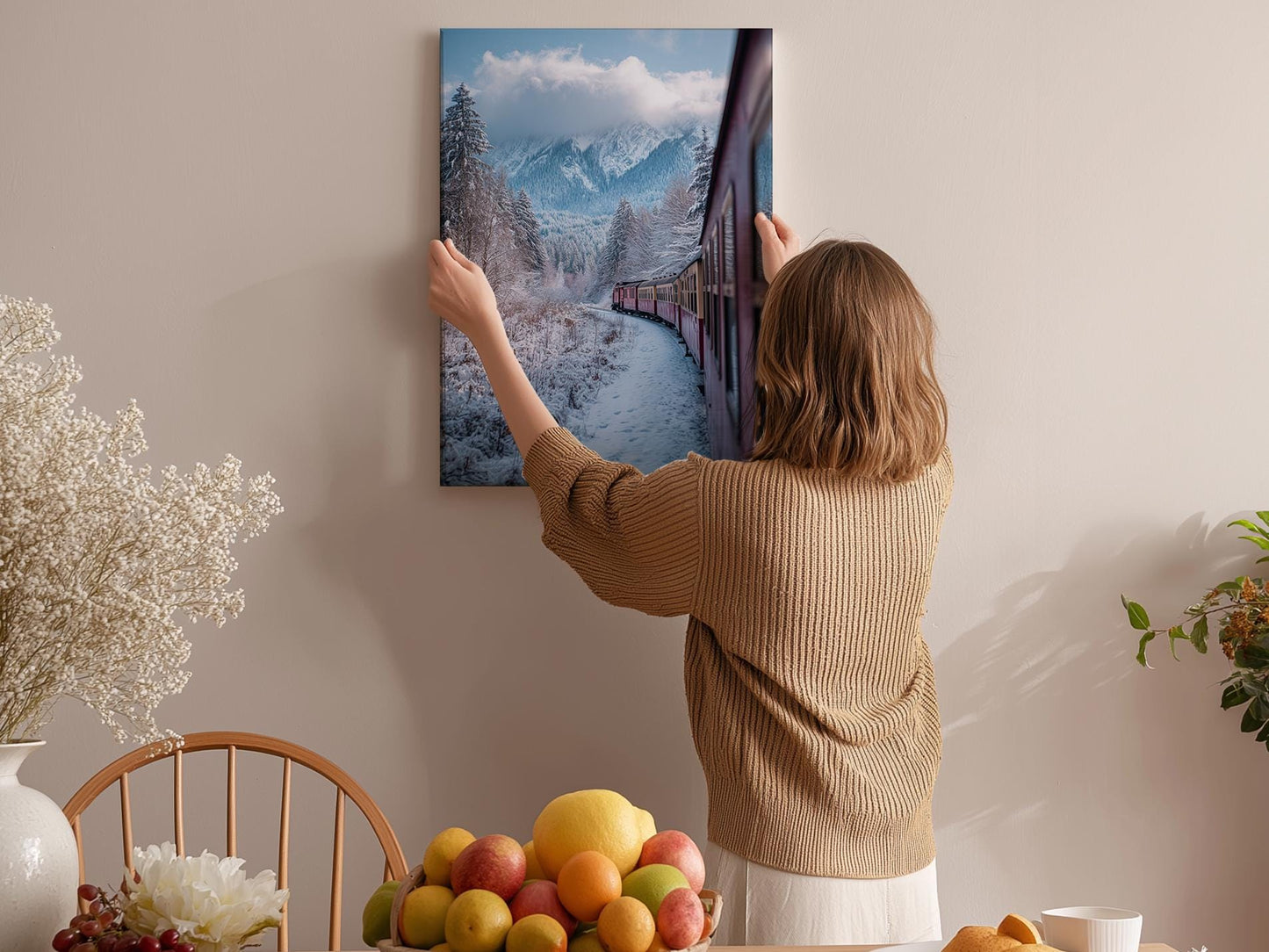 A woman hangs a framed picture of a snowy mountain landscape on a wall in a cozy room with a dining table, chairs, and various fruits.