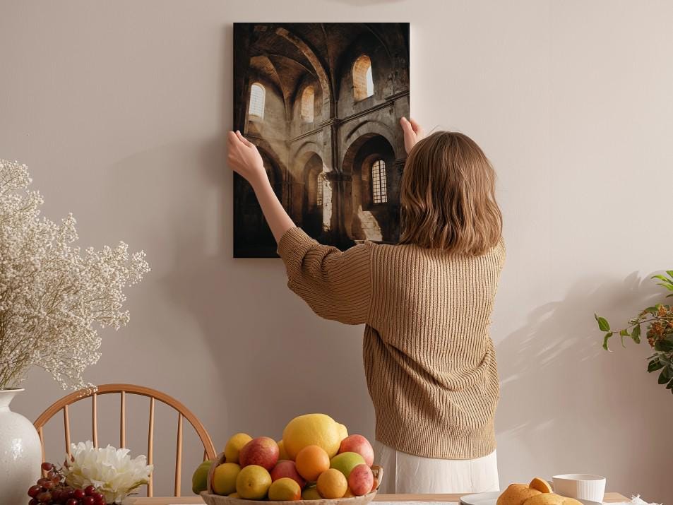 A woman hangs a framed picture on a wall in a room with a dining table, chairs, and various fruits and flowers.
