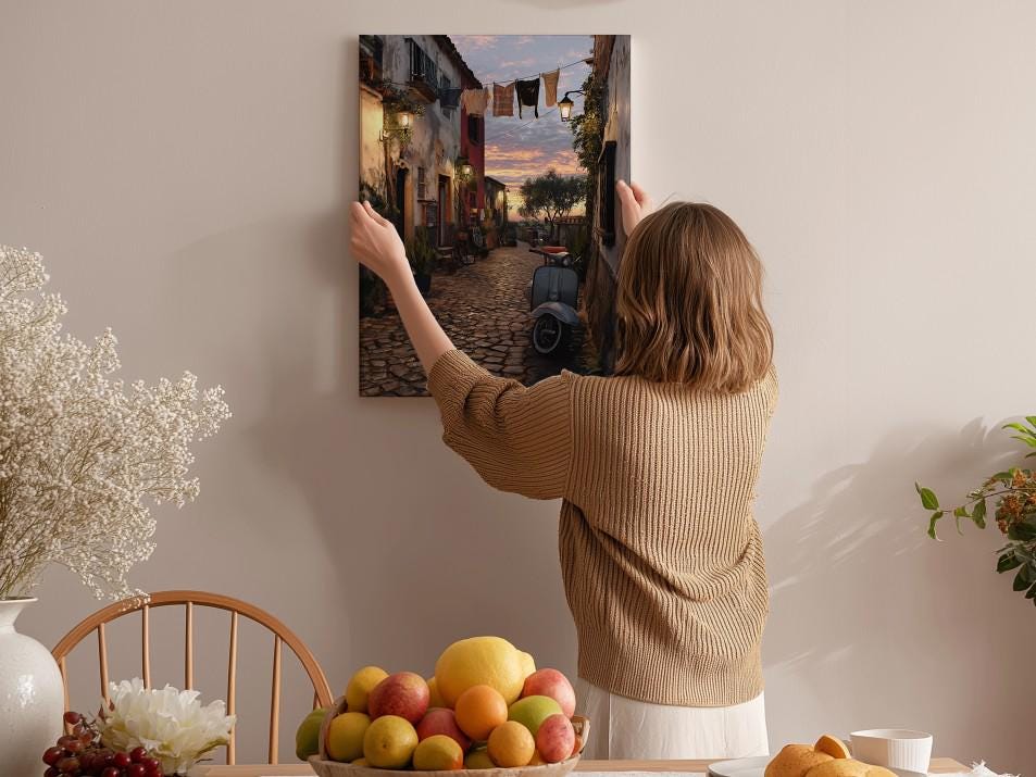 A woman hangs a framed picture of a cobblestone street with a sunset in the background on a wall in a cozy, homely setting.