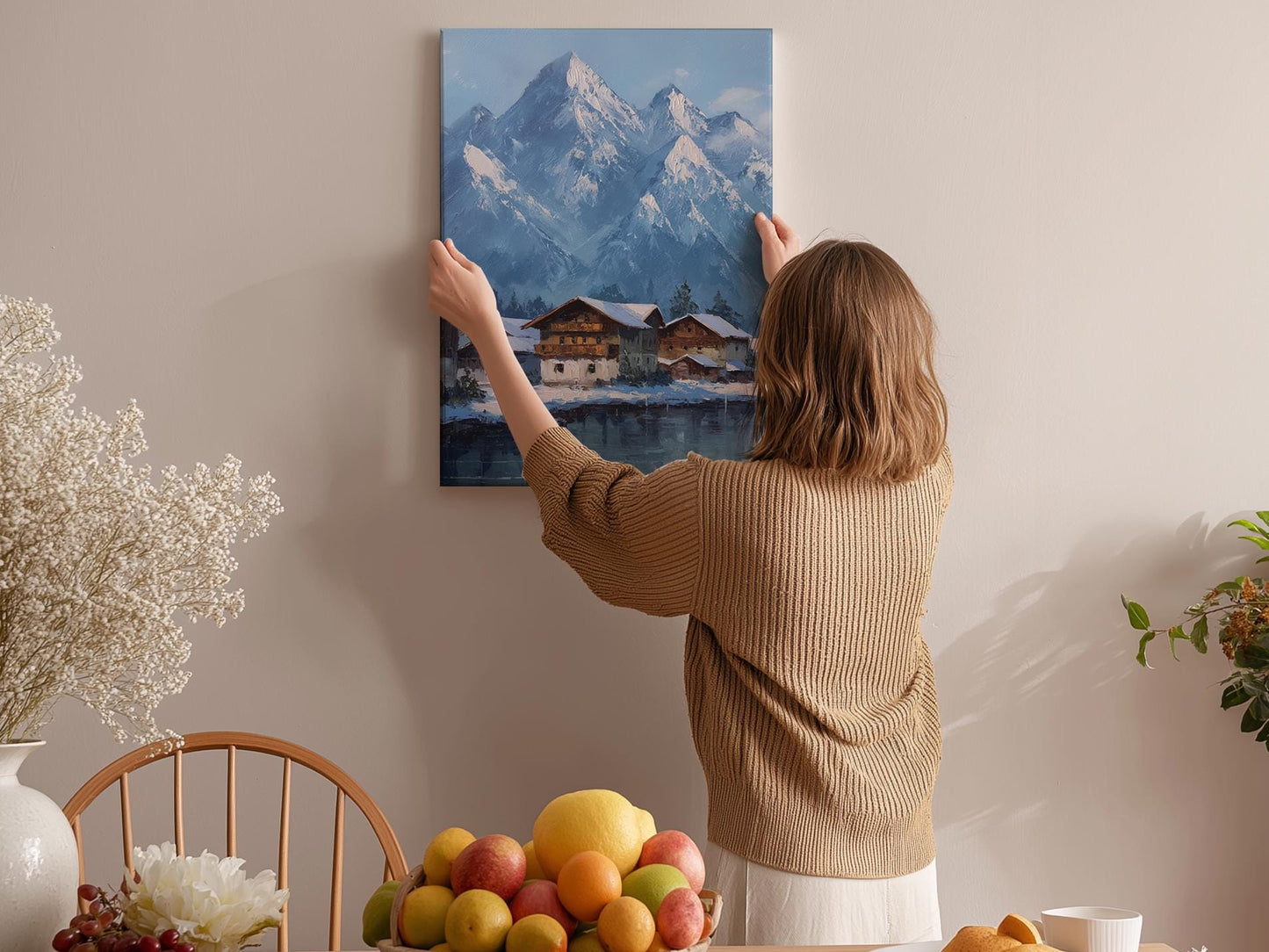 A woman hangs a painting of a mountain landscape on a wall in a cozy room with a dining table, chairs, and various decorative items.