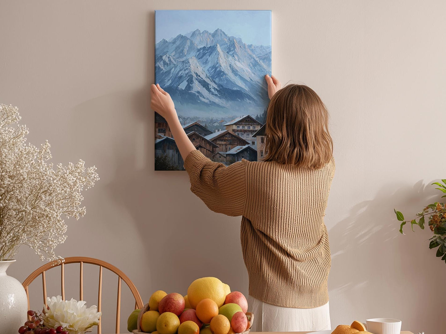 A woman hangs a painting of a mountain village on a wall in a cozy room with a dining table, chairs, and various fruits.