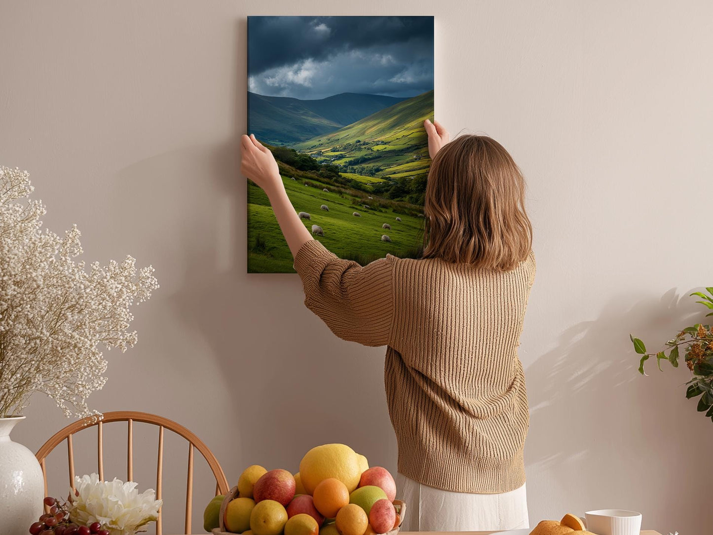 A woman hangs a landscape painting on a wall, which depicts a countryside scene with rolling hills and a valley.