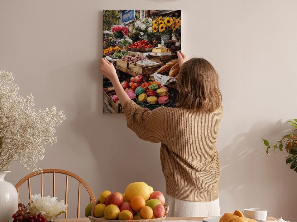 A woman hangs a framed photograph of a fruit stand on a wall in a room with a dining table, chairs, and various decorative items.