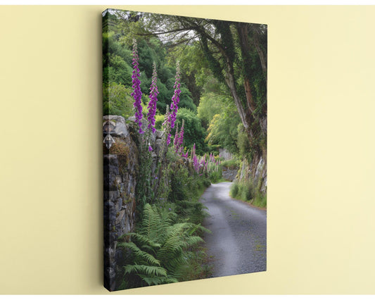 A stone wall lined with purple flowers and lush greenery leads down a winding path through a forested area.