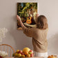A woman hangs a framed painting of a guitar on a wall in a cozy room with a dining table, chairs, and various fruits.