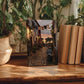 a wooden table with a framed photograph of a cobblestone street in a village, surrounded by potted plants and books.