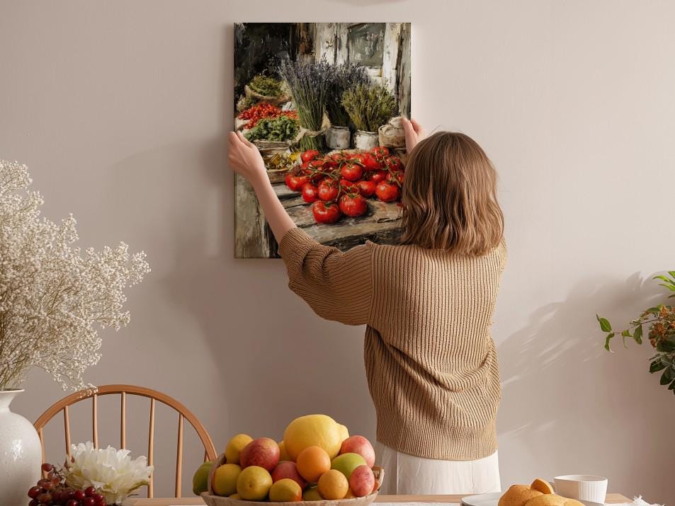 A woman hangs a framed painting of a fruit stand on a wall in a cozy kitchen setting.