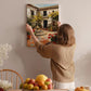 A woman hangs a framed picture of a Spanish-style house on a wall in a cozy, homely setting.