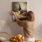 A woman hangs a painting of a table with wine, cheese, and fruit on a wall in a cozy, homely setting.