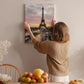 A woman hangs a framed picture of the Eiffel Tower on a wall in a room with a dining table, chairs, and various fruits.