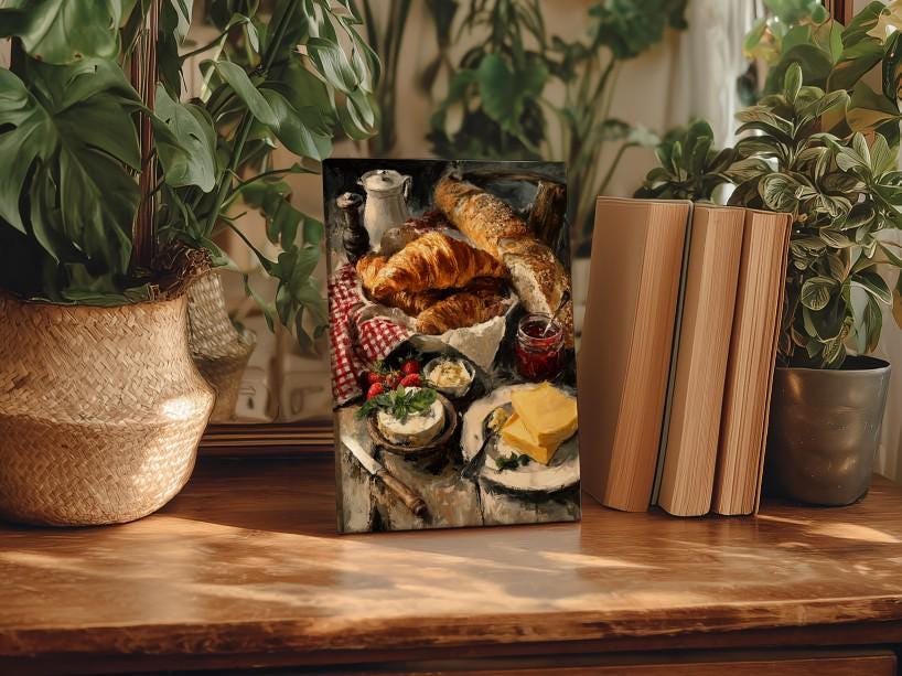 a wooden table with a framed painting of a breakfast scene, including croissants, cheese, and other food items. The painting is surrounded by potted plants and books, creating a cozy and inviting atmosphere.