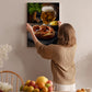 A woman hangs a framed photograph of a pretzel and a mug of beer on a wall in a cozy kitchen setting.