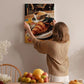A woman hangs a framed photograph of a croissant and coffee on a wall in a cozy kitchen setting.