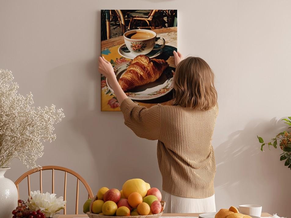 A woman hangs a framed photograph of a croissant and coffee on a wall in a cozy kitchen setting.