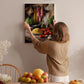 A woman hangs a framed picture of a kitchen scene on a wall, surrounded by various fruits and vegetables.