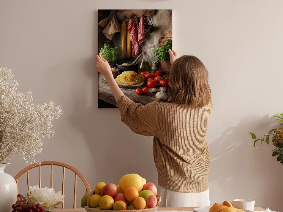 A woman hangs a framed picture of a kitchen scene on a wall, surrounded by various fruits and vegetables.
