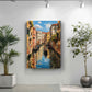 a painting of a canal in Venice, Italy, with colorful buildings lining the waterway and a gondola visible in the distance. The painting is hung on a white wall, and there are two potted plants in the foreground, one on the left and one on the right.