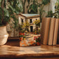 a wooden table with a framed painting of a courtyard scene, surrounded by potted plants and books.
