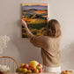 A woman hangs a landscape painting on a wall, which depicts a countryside scene with rolling hills, vineyards, and trees.