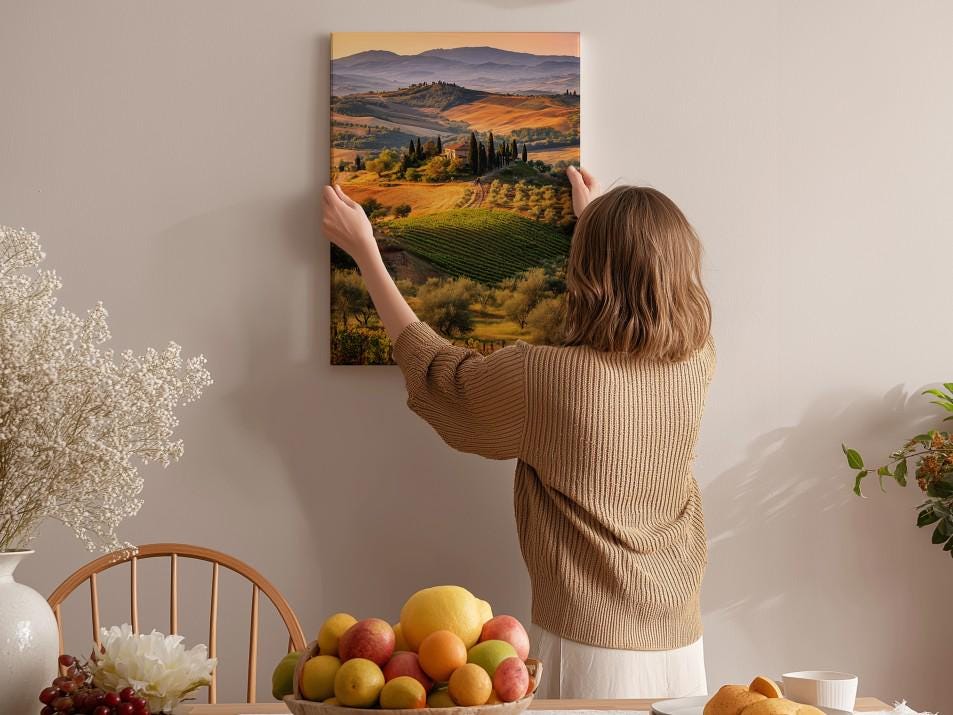 A woman hangs a landscape painting on a wall, which depicts a countryside scene with rolling hills, vineyards, and trees.
