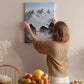 A woman hangs a framed picture of a snowy mountain landscape on a wall in a cozy room with a dining table, chairs, and various fruits.