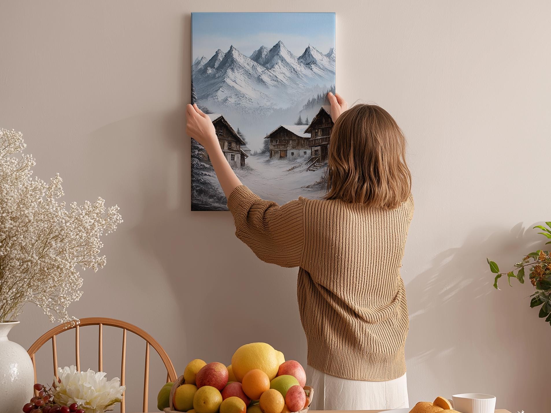 A woman hangs a framed picture of a snowy mountain landscape on a wall in a cozy room with a dining table, chairs, and various fruits.