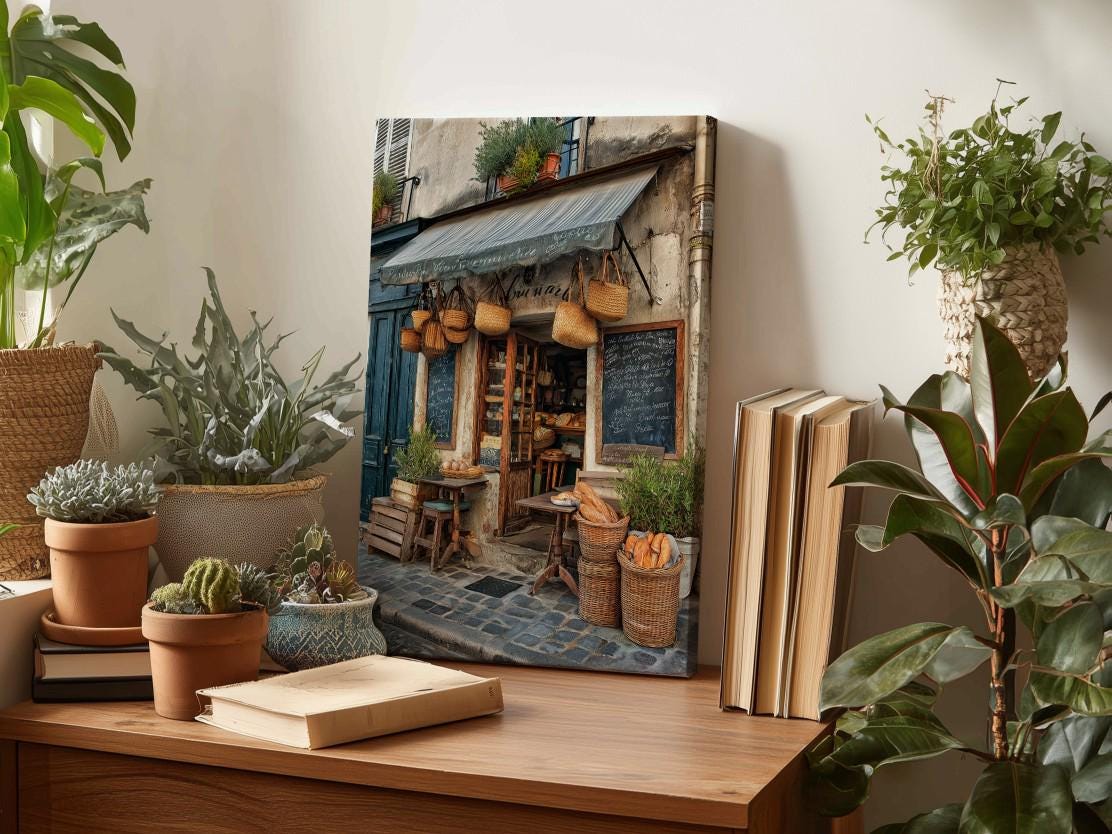 a wooden table with a painting of a quaint bakery shop displayed on it. The table also has several potted plants, books, and other decorative items.