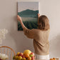 A woman hangs a landscape painting on a wall, surrounded by a table with fruit and flowers, creating a cozy and inviting atmosphere.