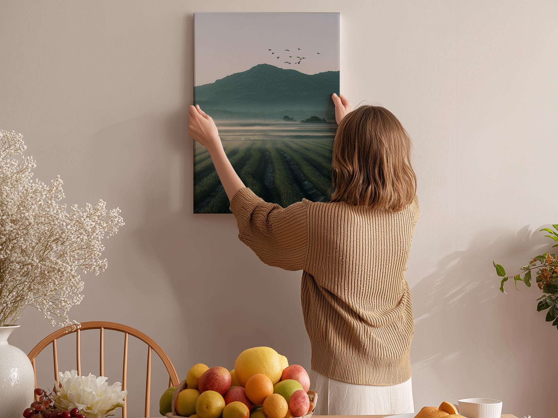 A woman hangs a landscape painting on a wall, surrounded by a table with fruit and flowers, creating a cozy and inviting atmosphere.