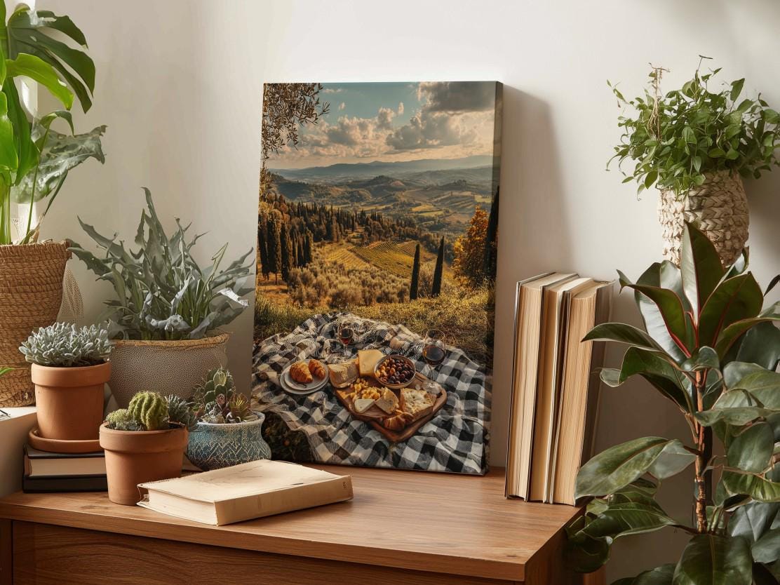 a wooden table with a checkered tablecloth, a picnic basket filled with food, and several potted plants. In the background, there is a framed landscape photograph depicting a scenic countryside view.