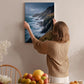 A woman hangs a framed picture of a coastal landscape on a wall in a room with a dining table, chairs, and various fruits.