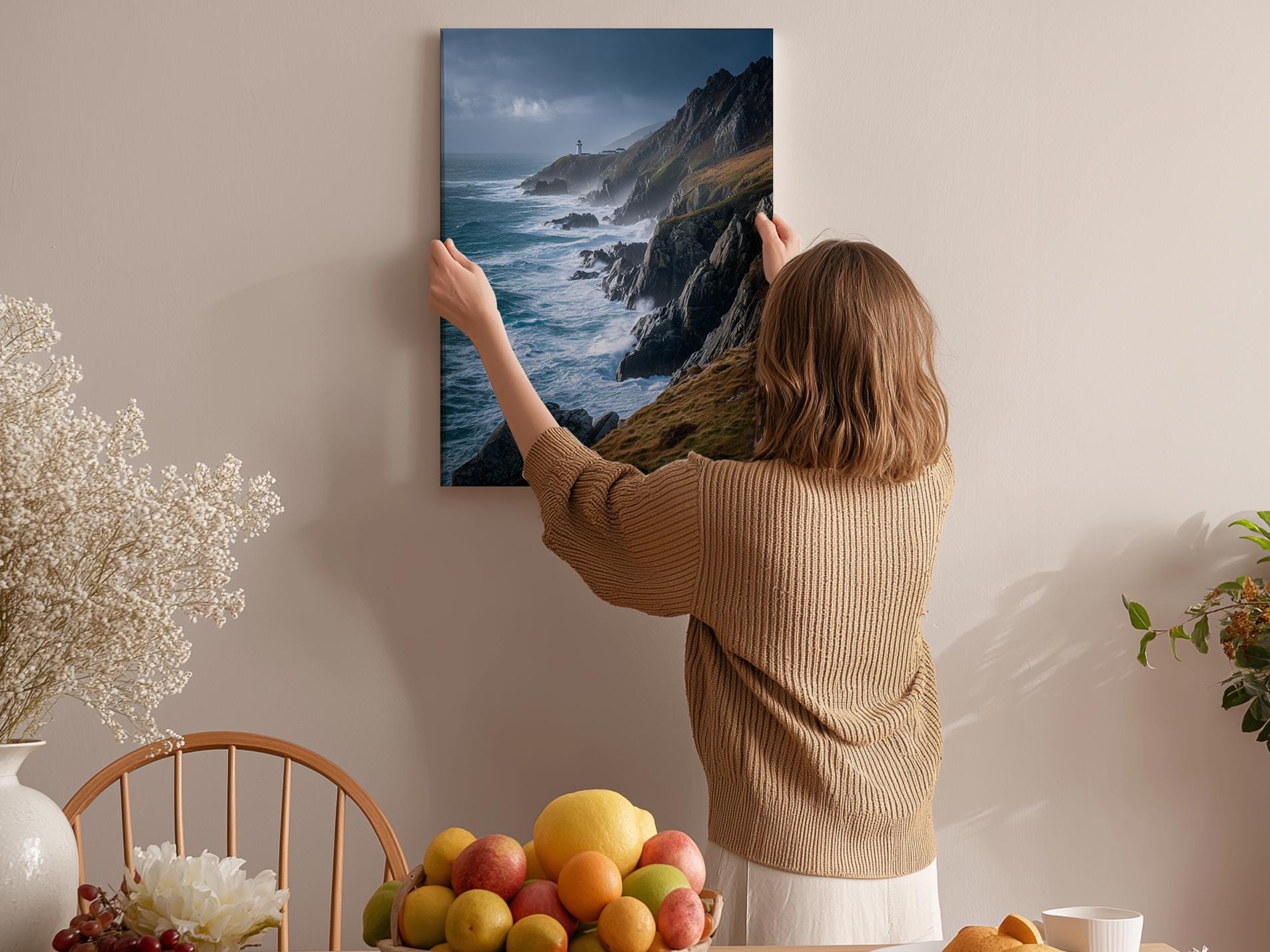 A woman hangs a framed picture of a coastal landscape on a wall in a room with a dining table, chairs, and various fruits.