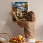 A woman hangs a painting of a house on a wall in a room with a dining table, chairs, and various fruits and flowers.