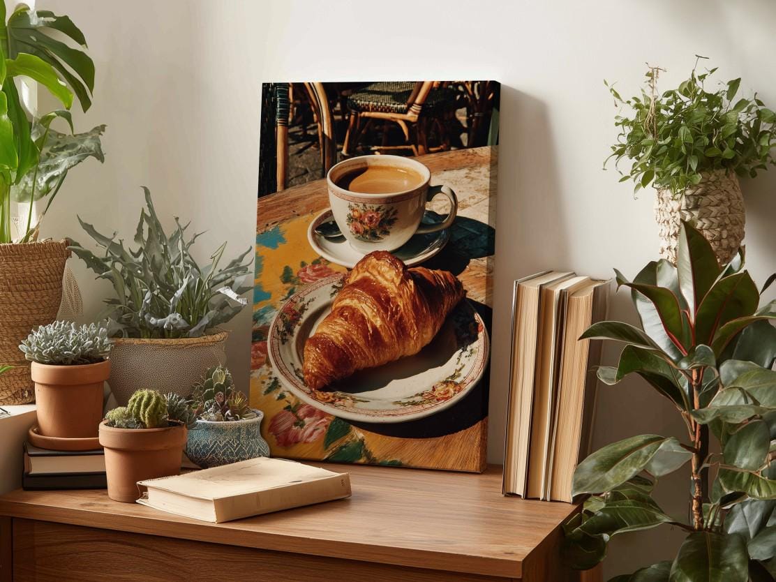 a wooden table with a croissant on a plate, surrounded by potted plants and a book. In the background, there is a framed photograph of a cup of coffee and a croissant on a table.