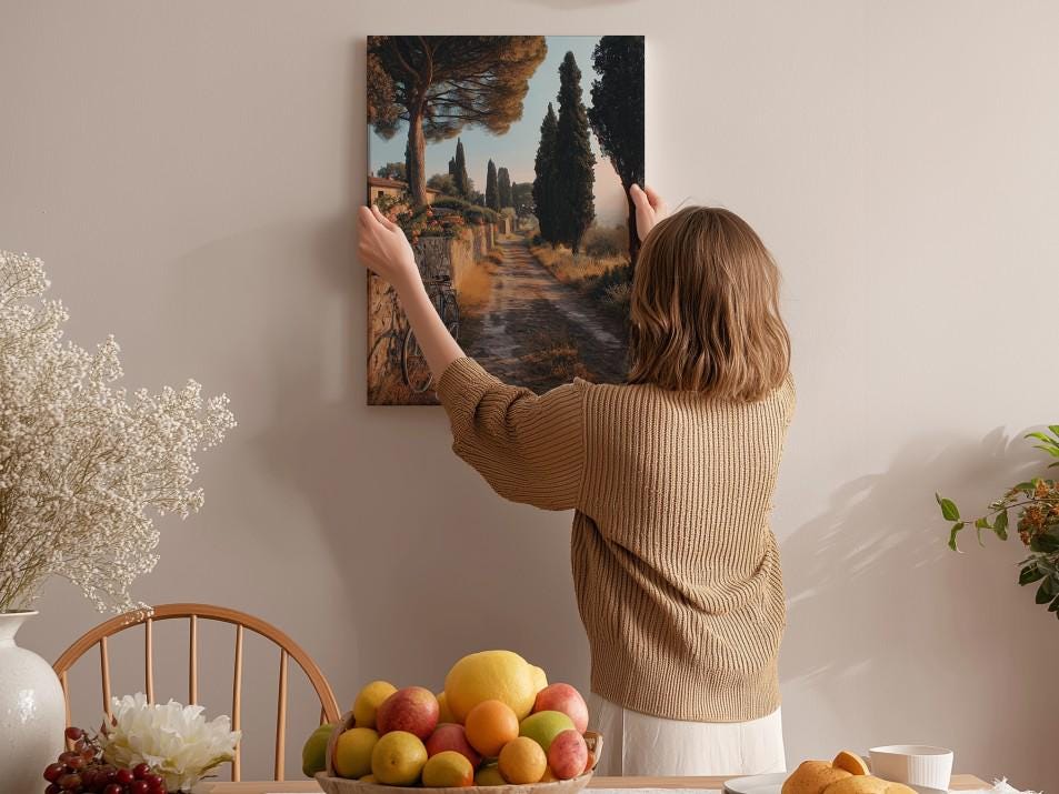 A woman hangs a framed picture of a scenic landscape on a wall in a cozy room with a dining table, chairs, and various decorative items.