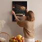 A woman hangs a framed picture on a wall in a cozy, homely setting with a dining table, chairs, and various decorative elements.