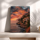 a coastal landscape with a house perched on a cliff overlooking the ocean. The sky is filled with vibrant colors, suggesting a stunning sunset or sunrise. The photograph is framed and displayed on a shelf, with two vases placed next to it.