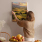 A woman hangs a landscape painting on a wall, surrounded by a dining table with fruit and flowers, creating a cozy and inviting atmosphere.