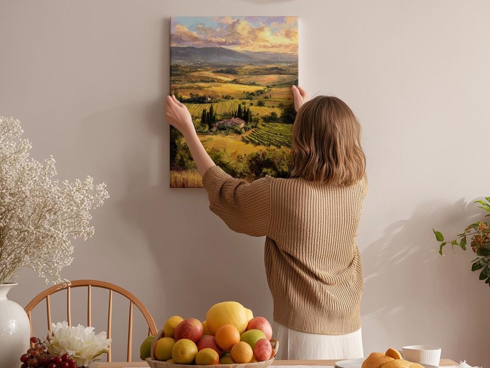 A woman hangs a landscape painting on a wall, surrounded by a dining table with fruit and flowers, creating a cozy and inviting atmosphere.